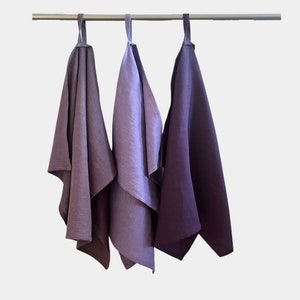 May include: Three linen kitchen towels hanging on a rod. The towels are a gradient of purple shades, from light to dark.