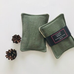 May include: Two green kitchen scrubbers with white edging. The scrubber on the right has a black label that reads "Cool & Mellow Reusable Kitchen Sponge 100% linen".