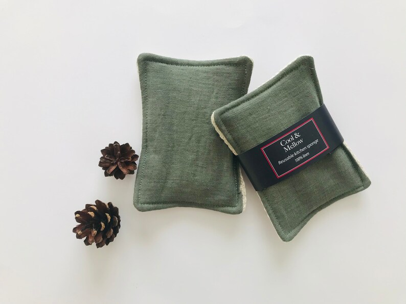May include: Two green kitchen scrubbers with white edging. The scrubber on the right has a black label that reads "Cool & Mellow Reusable Kitchen Sponge 100% linen".