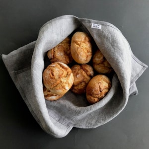 Op de afbeelding: Een grijze linnen broodmand gevuld met acht ronde, goudbruine broodjes. De mand heeft een klein wit label met de tekst "Cook & Moore" erop.