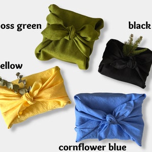 May include: Four fabric-wrapped gifts in different colors: moss green, yellow, black, and cornflower blue. Each gift is tied with a knot and decorated with a sprig of greenery.