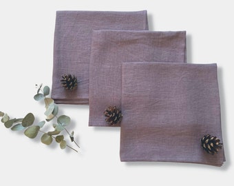May include: Three square mauve-coloured fabric napkins with a visible woven texture. Each napkin is folded into a square shape and arranged in a staggered stack. Three small brown pinecones are placed on the napkins, and a sprig of eucalyptus leaves.