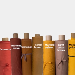 Op de afbeelding: Tien rollen stof in verschillende tinten oranje, bruin en geel. De stofrollen zijn gelabeld met de kleuren: Oranje, Terracotta, Hazelnootbruin, Kaneel, Kameelbruin, Mosterdgeel, Licht cacao bruin, Mangogeel en Geel.