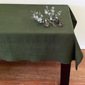 Pode incluir: Uma toalha de mesa de linho verde escuro sobre uma mesa. A toalha de mesa é de cor sólida e tem uma aparência ligeiramente texturizada. Um ramo de eucalipto e pinhas são dispostos sobre a toalha de mesa.