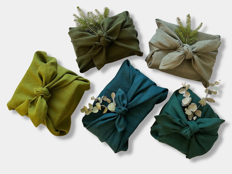 May include: Five fabric gift wraps in various shades of green, including olive, forest green, and teal. Each wrap is tied with a knot and decorated with sprigs of greenery.