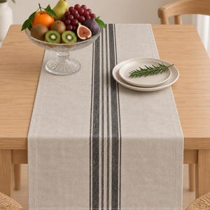 May include: A rectangular beige table runner featuring a central vertical pattern of three dark charcoal gray stripes. The runner is laid flat on a light wood table, topped with a glass fruit bowl containing assorted fresh fruit and a small white plate.