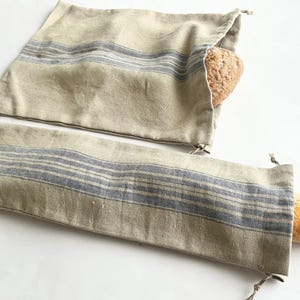 Puede incluir: Dos bolsas de pan de lino beige con rayas azules. Una bolsa está llena de una hogaza de pan, la otra está llena de una baguette.