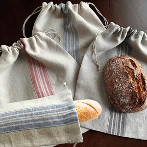 May include: Three linen bread bags with striped patterns. One bag holds a loaf of bread, and another holds a baguette.