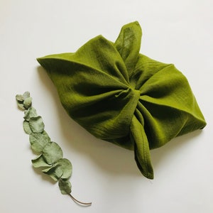 Puede incluir: Una bolsa de tela verde atada con un nudo, sobre un fondo blanco, con una rama de hojas de eucalipto secas a la izquierda.