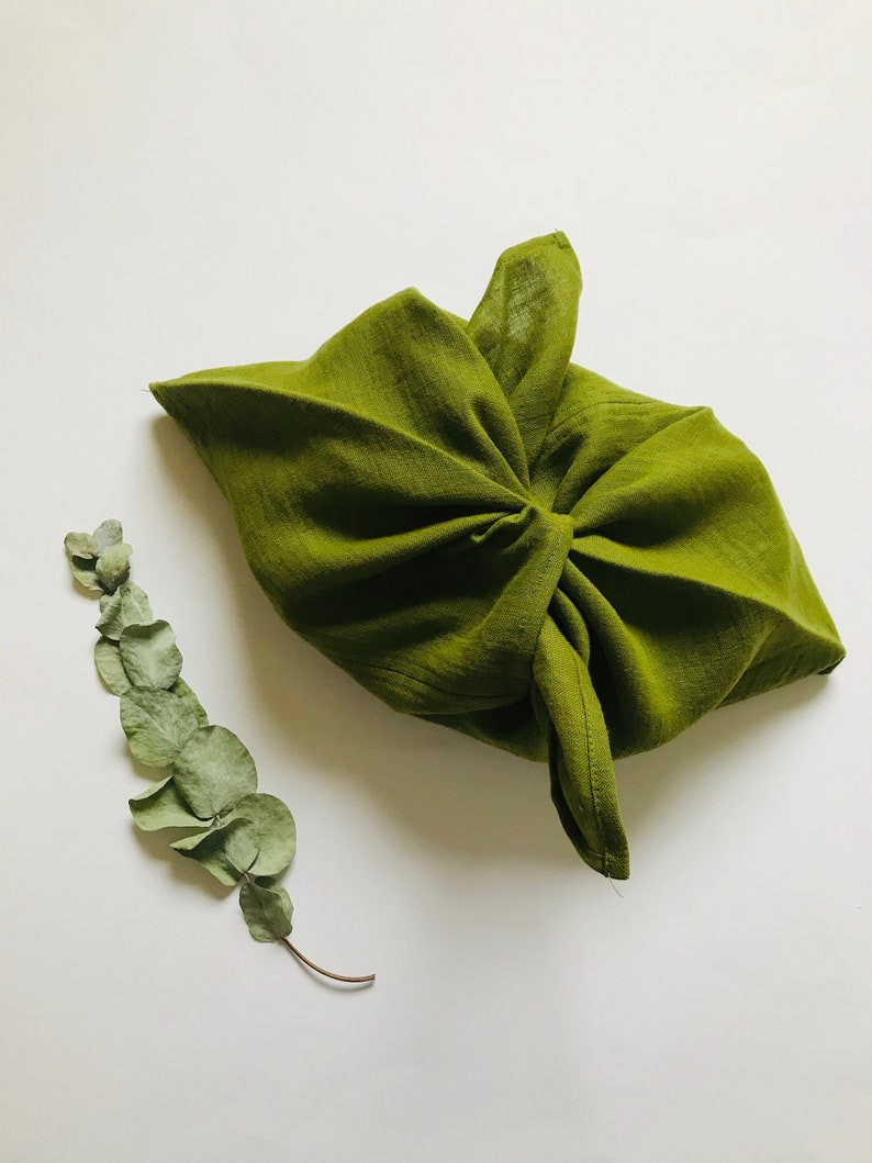 Puede incluir: Una bolsa de tela verde atada con un nudo, sobre un fondo blanco, con una rama de hojas de eucalipto secas a la izquierda.