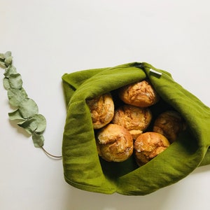 Puede incluir: Una bolsa de tela verde llena de panecillos redondos de color marr&oacute;n dorado. La bolsa est&aacute; doblada sobre la parte superior de los panecillos, y los bordes de la bolsa son visibles.