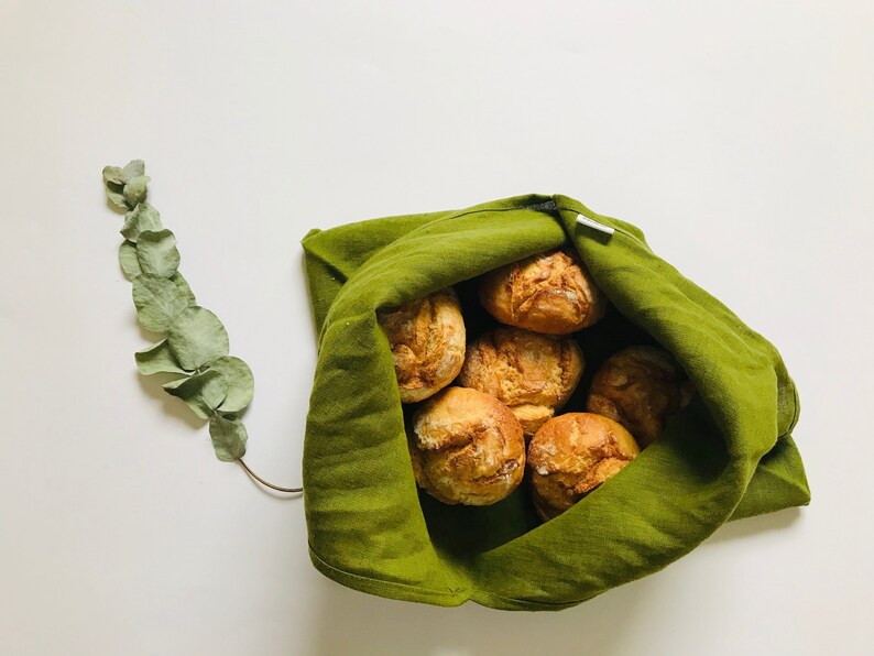 Puede incluir: Una bolsa de tela verde llena de panecillos redondos de color marr&oacute;n dorado. La bolsa est&aacute; doblada sobre la parte superior de los panecillos, y los bordes de la bolsa son visibles.