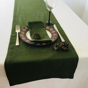 May include: A dark green linen table runner with a matching napkin and place setting. The plate has a brown and white spiral design. A fork, knife, and wine glass are also present. Two small pine cones are placed on the table.