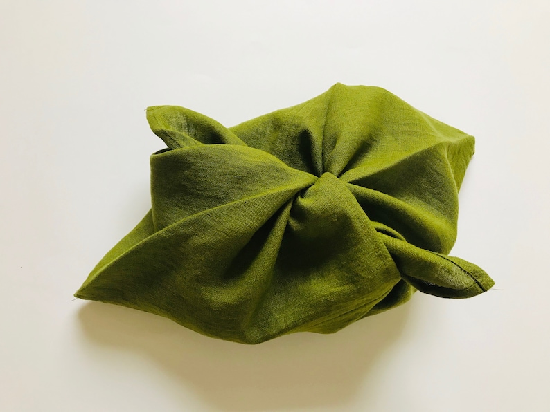 Puede incluir: Una bolsa de tela verde doblada en forma de cuadrado. La tela es texturizada y tiene un ligero brillo.
