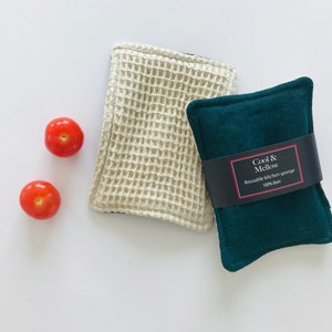May include: Two reusable kitchen sponges, one white and one dark green. The green sponge has a label that reads "Cool & Mellow Reusable kitchen sponge 100% linen". Two red tomatoes are in the background.