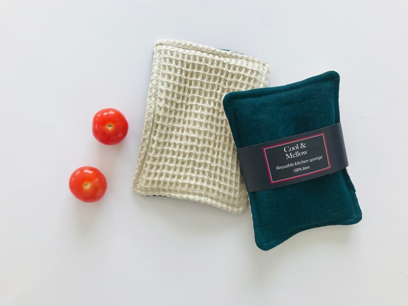 May include: Two reusable kitchen sponges, one white and one dark green. The green sponge has a label that reads "Cool & Mellow Reusable kitchen sponge 100% linen". Two red tomatoes are in the background.