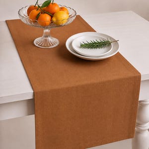 Puede incluir: Un camino de mesa rectangular de color óxido sólido con un borde cosido. Un cuenco de cristal transparente sobre un pedestal contiene varias naranjas y un limón. Junto a él, dos platos de cerámica blanca apilados tienen una rama de romero.
