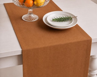May include: A solid rust-colored rectangular table runner with a stitched border. A clear glass pedestal bowl holds several oranges and a lemon. Next to it, two stacked white ceramic plates are topped with a single sprig of green rosemary.