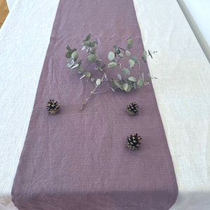 May include: A purple linen table runner with three pine cones and a sprig of eucalyptus leaves on a white linen tablecloth.