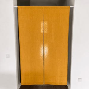 Può includere: Una tenda da porta in tessuto color giallo senape tinta unita appesa a un bastone a pressione bianco. La tenda è divisa al centro in due pannelli verticali che coprono un passaggio tra due pareti bianche. Il tessuto ha una trama visibile.
