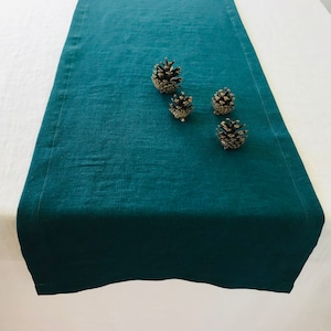 May include: A teal linen table runner with four small pine cones on top.