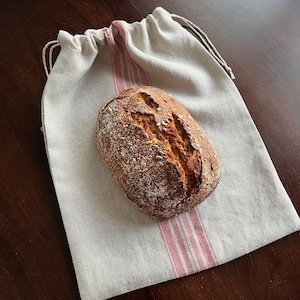 May include: A loaf of sourdough bread with sunflower seeds in a white linen bag with red stripes.