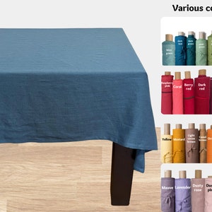 Könnte beinhalten: Eine blaue Leinen-Tischdecke auf einem Holztisch. Das Bild zeigt verschiedene Farben von Stoffrollen, darunter Himbeerrosa, Korallenrot, Beerrot, Dunkelrot, Pflaumenlila, Dunkelviolett, Gelb, Senf, helles Kakaobraun, Kamelbraun, Orange, Terrakotta, Haselnuss-Karamellbraun, Mauve, Lavendel, staubiges Rosa, staubiges Rosa, helles Pfirsichrosa und Vanille.