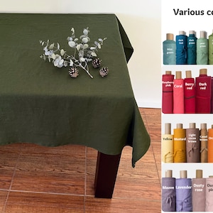 May include: A green linen tablecloth with a sprig of eucalyptus and pine cones on top. The tablecloth is shown on a wooden table. A sample card shows various colors of linen fabric, including raspberry pink, coral, berry red, dark red, plum purple, dark purple, yellow, mustard, light cocoa brown, camel, orange, terracotta, hazelnut brown, mauve, lavender, dusty rose, dusty pink, light peachy pink, and vanilla.