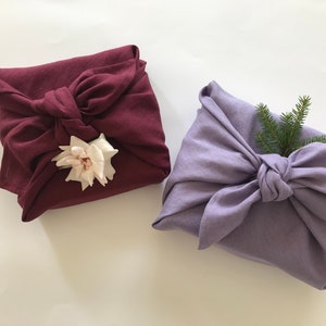 May include: Two fabric-wrapped gifts, one in a deep burgundy color with a white flower, and the other in a light lavender color with a sprig of green pine.