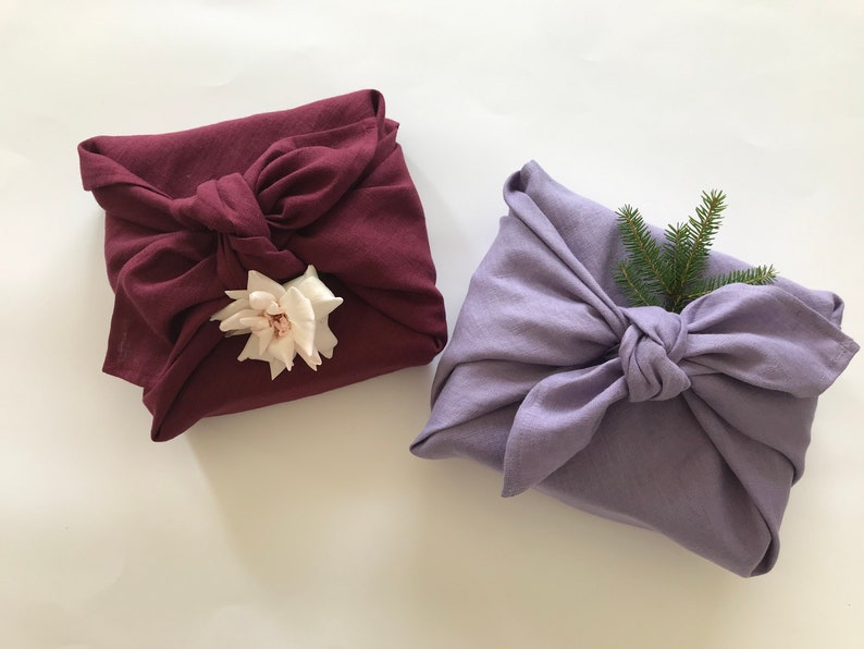 May include: Two fabric-wrapped gifts, one in a deep burgundy color with a white flower, and the other in a light lavender color with a sprig of green pine.