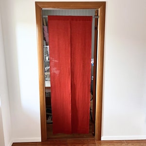 May include: A doorway with a wooden frame and a vibrant red curtain hanging in the opening. The curtain is made of a textured fabric and is split into two panels. The walls are white, and the floor is wood.
