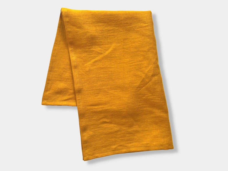 K&ouml;nnte beinhalten: Ein rechteckiges St&uuml;ck Stoff in einer einfarbigen senfgelben Farbe. Das Textil weist eine feine, gewebte Struktur mit sichtbaren horizontalen Maserungslinien und weichen Falten im gesamten Material auf, was ein schlichtes Erscheinungsbild erzeugt.