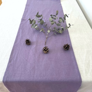 May include: A lilac linen table runner on a white tablecloth, decorated with a sprig of eucalyptus and three small pine cones. The runner adds colour and texture to the neutral table setting.