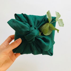 May include: A green fabric gift wrap with a knot tied in the center. A sprig of green leaves is placed on top of the knot.