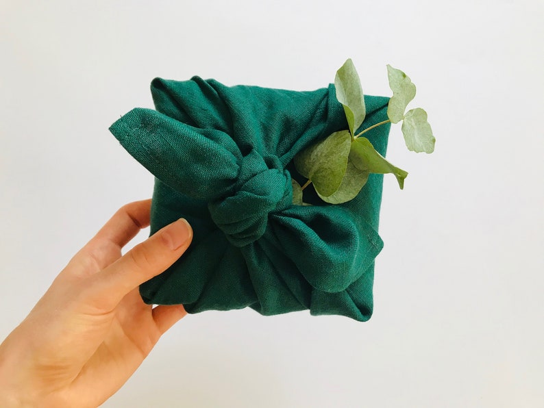May include: A green fabric gift wrap with a knot tied in the center. A sprig of green leaves is placed on top of the knot.