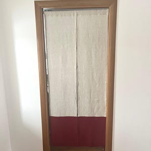 May include: A doorway curtain with a natural linen upper section and a deep red lower section. The curtain hangs within a wooden door frame, creating a simple yet stylish room divider or doorway accent.