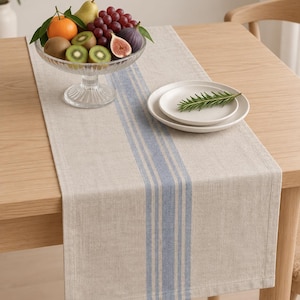 May include: A rectangular table runner made of light beige fabric featuring a central vertical pattern of three distinct blue stripes. The runner is laid flat across a wooden table, providing a neutral base for a glass fruit bowl and plates.