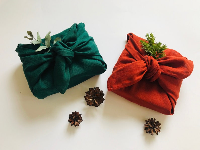 May include: Two fabric-wrapped gifts, one green and one red, with sprigs of greenery on top. The gifts are surrounded by small brown pine cones.