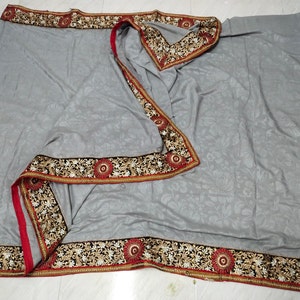 May include: A gray textile with a floral pattern and ornate borders. The borders feature intricate gold and black embroidery with red accents. The fabric is draped, showcasing the detailed design and craftsmanship.