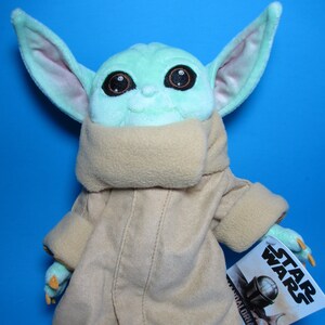 May include: A green plush toy of a character from the Star Wars franchise, The Mandalorian. The toy is wearing a brown hooded robe and has large ears. The toy is holding a tag that says "Star Wars".