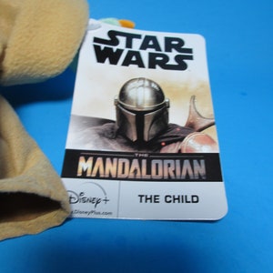 May include: A Star Wars The Mandalorian plush toy with a tag featuring the Mandalorian character in his helmet and the text "The Mandalorian" and "The Child". The tag also includes the Disney+ logo.