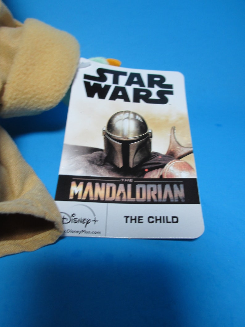 May include: A Star Wars The Mandalorian plush toy with a tag featuring the Mandalorian character in his helmet and the text "The Mandalorian" and "The Child". The tag also includes the Disney+ logo.