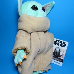 May include: A plush toy of The Child, also known as Baby Yoda, from the Star Wars series The Mandalorian. The toy is wearing a brown robe and has large green ears and big brown eyes. The toy is standing on a blue background and has a Star Wars tag attached to it.
