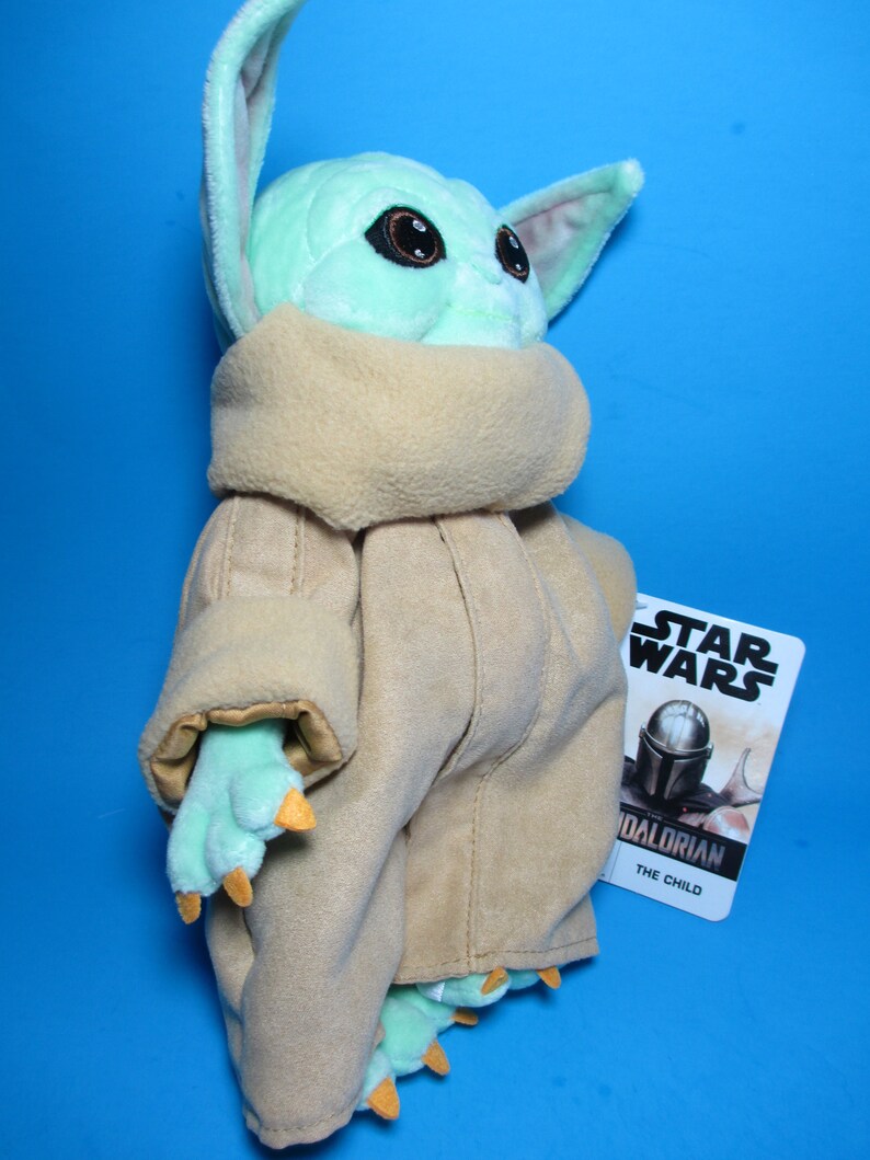 May include: A plush toy of The Child, also known as Baby Yoda, from the Star Wars series The Mandalorian. The toy is wearing a brown robe and has large green ears and big brown eyes. The toy is standing on a blue background and has a Star Wars tag attached to it.