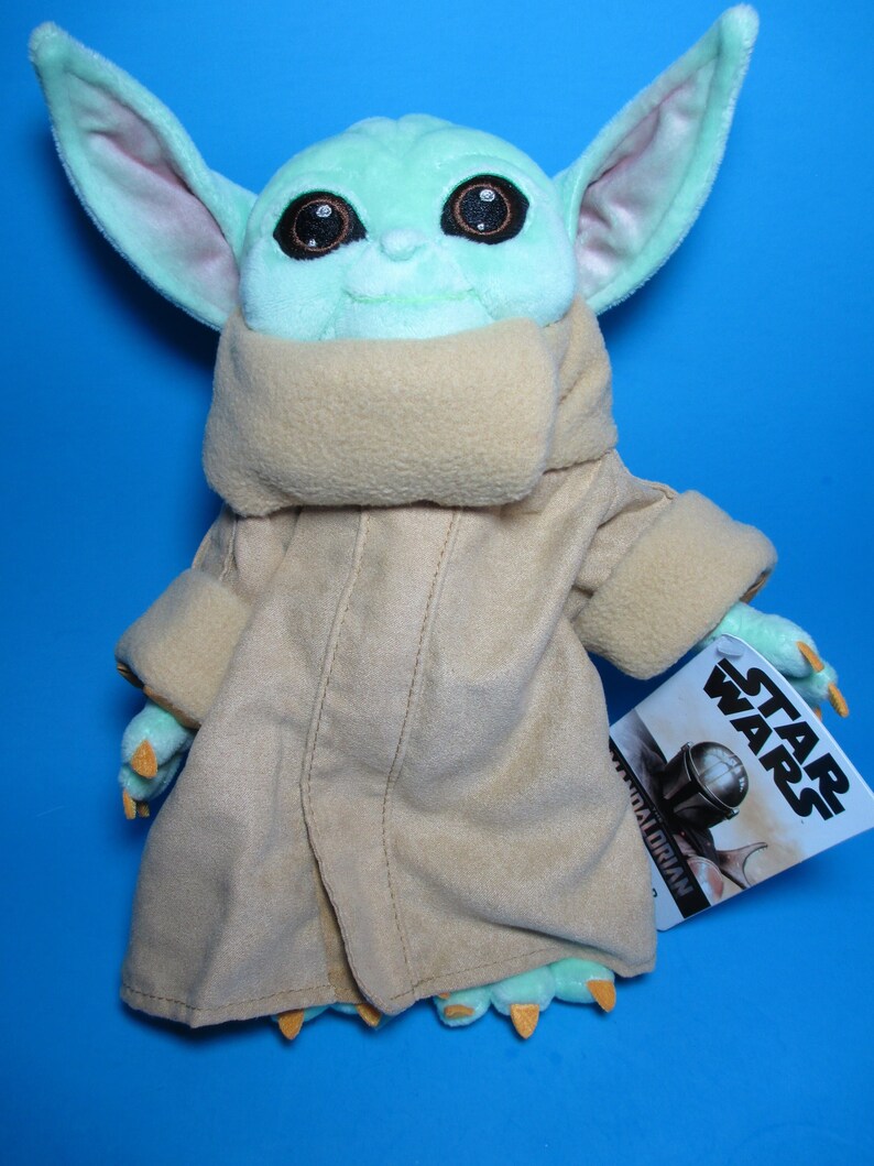 May include: A green plush toy of a character from the Star Wars franchise, The Mandalorian. The toy is wearing a brown hooded robe and has large ears. The toy is holding a tag that says "Star Wars".
