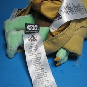 May include: A green and brown plush toy of Yoda from Star Wars. The toy is wearing a brown robe and has a white tag with the Disney logo and the text "shopDisney.com".