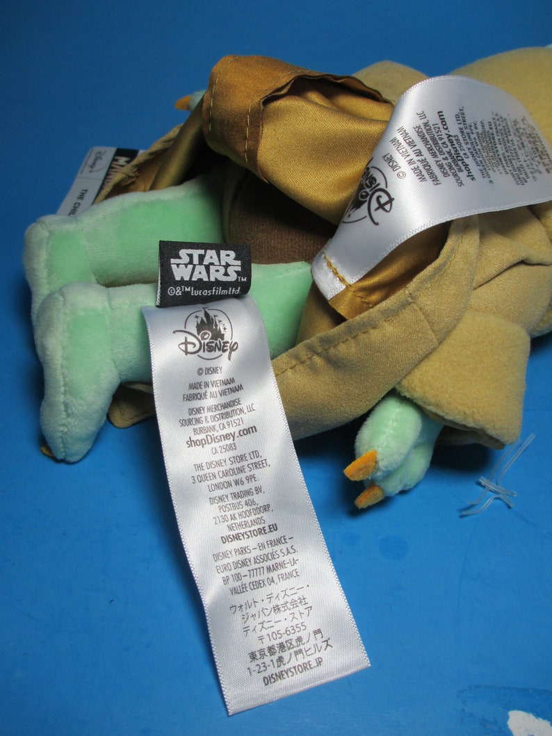 May include: A green and brown plush toy of Yoda from Star Wars. The toy is wearing a brown robe and has a white tag with the Disney logo and the text "shopDisney.com".