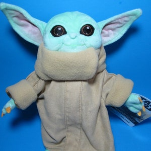 May include: A green plush toy of the character Baby Yoda from the Star Wars series, wearing a brown hooded robe. The toy has large, dark eyes and a small mouth. The toy is holding a tag that says "Star Wars The Mandalorian"
