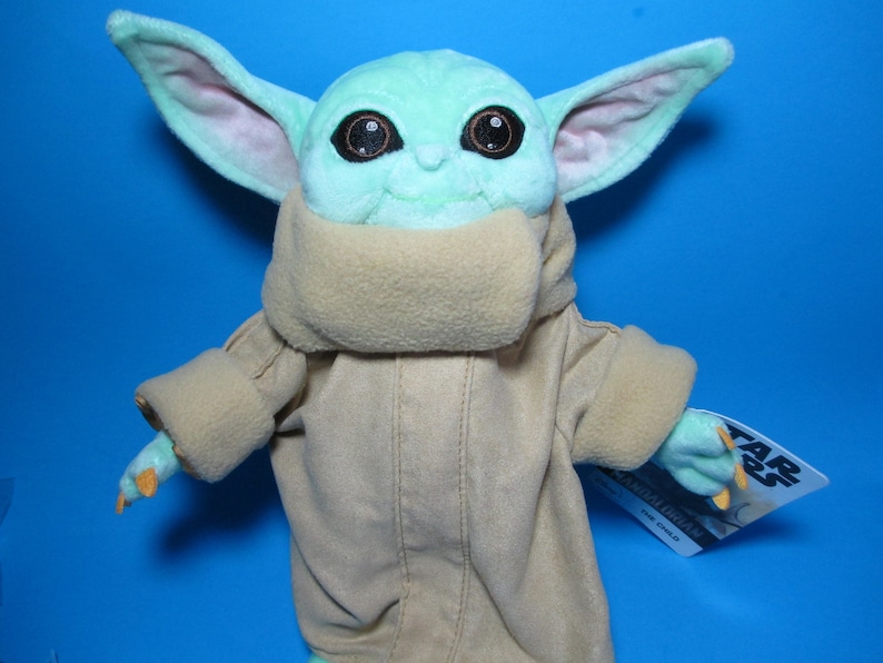 May include: A green plush toy of the character Baby Yoda from the Star Wars series, wearing a brown hooded robe. The toy has large, dark eyes and a small mouth. The toy is holding a tag that says "Star Wars The Mandalorian"