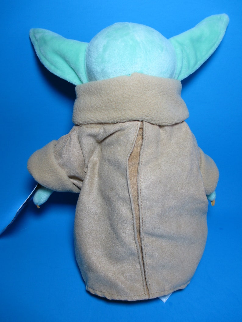 May include: A plush toy of a green character with large ears wearing a brown coat. The toy is facing away from the camera.